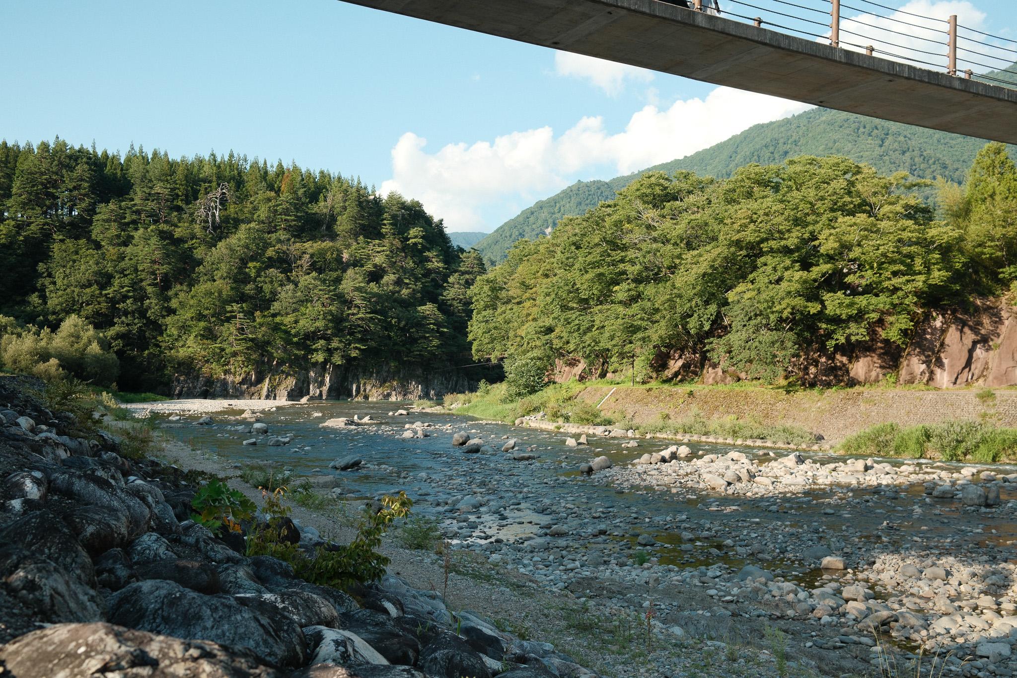 A wide, shallow river full of round grey stones, running between forested hills under a concrete footbridge on a sunny afternoon.